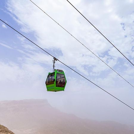 Tolip Resort El Galala Majestic Ajn Suchna Zewnętrze zdjęcie The photo shows a green cable car suspended in the air, traveling along overhead cables. The backdrop features a hazy landscape, likely mountainous or elevated terrain, with a cloudy sky. The scene suggests a scenic transportation method, possibly us