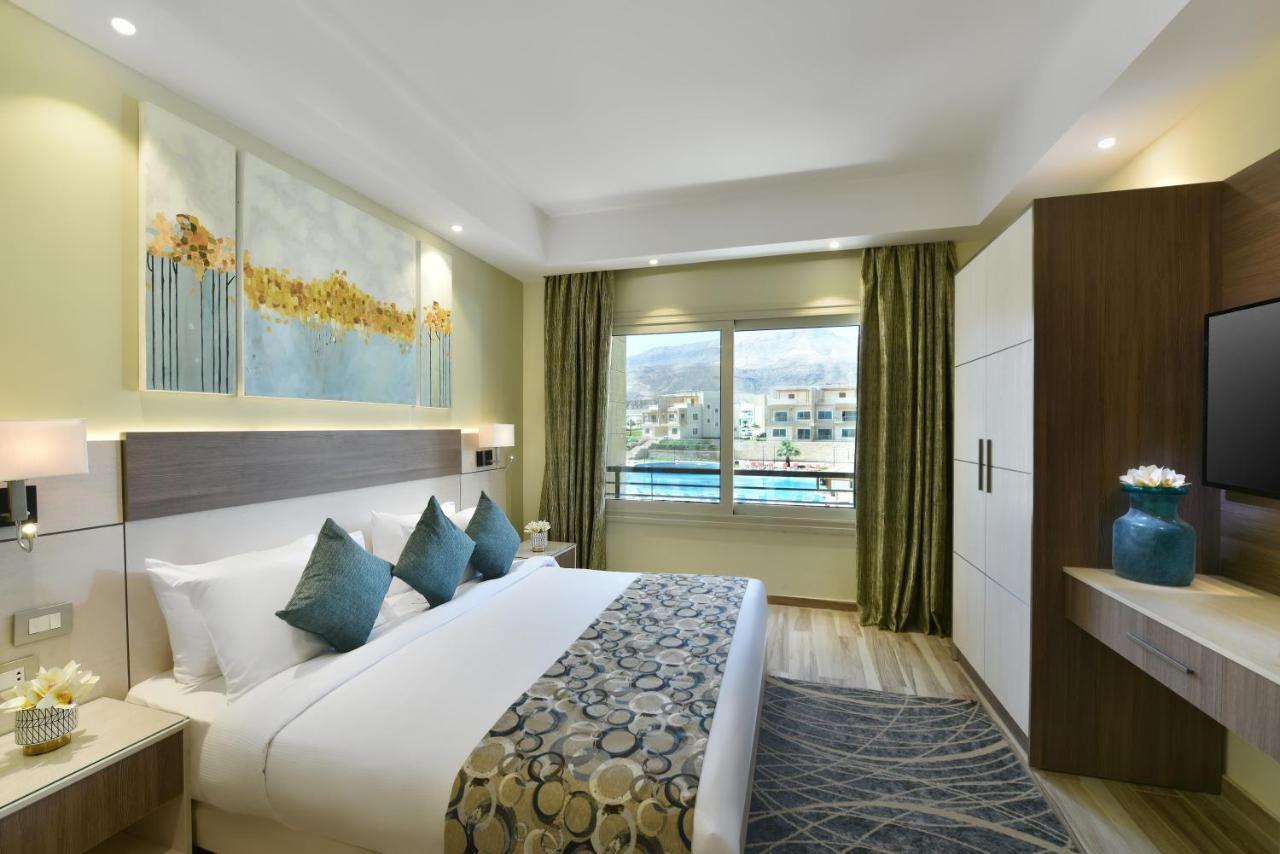 Tolip Resort El Galala Majestic Ajn Suchna Zewnętrze zdjęcie The photo shows a modern hotel room. It features a large bed with white bedding and several decorative pillows in shades of teal. Above the bed, there are two pieces of wall art with a landscape theme. The room has a large window with green curtains,