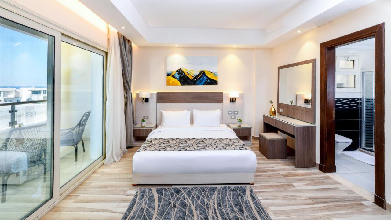 Tolip Resort El Galala Majestic Ajn Suchna Zewnętrze zdjęcie The photo shows a modern hotel room featuring a large bed with white linens and a decorative throw. The room has a neutral color palette, accented with warm lighting. There is a piece of artwork above the bed depicting mountains. To the left, there a