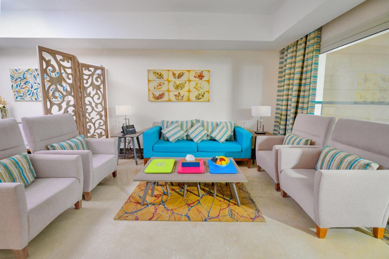 Tolip Resort El Galala Majestic Ajn Suchna Zewnętrze zdjęcie The photo shows a stylish and colorful living room. It features a bright blue sofa with patterned cushions, complemented by two light gray armchairs. In front of the sofa, there's a wooden coffee table with vibrant trays in pink, green, and yellow. A