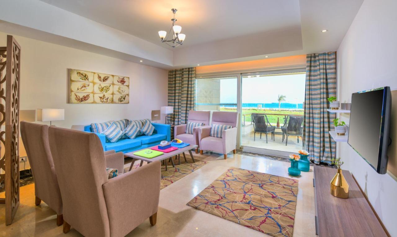 Tolip Resort El Galala Majestic Ajn Suchna Zewnętrze zdjęcie The photo shows a bright and modern living room. It features a blue sofa with decorative cushions, two light-colored armchairs, and a coffee table. A large area rug adds warmth to the space, which is decorated with a subtle pattern. The walls have so