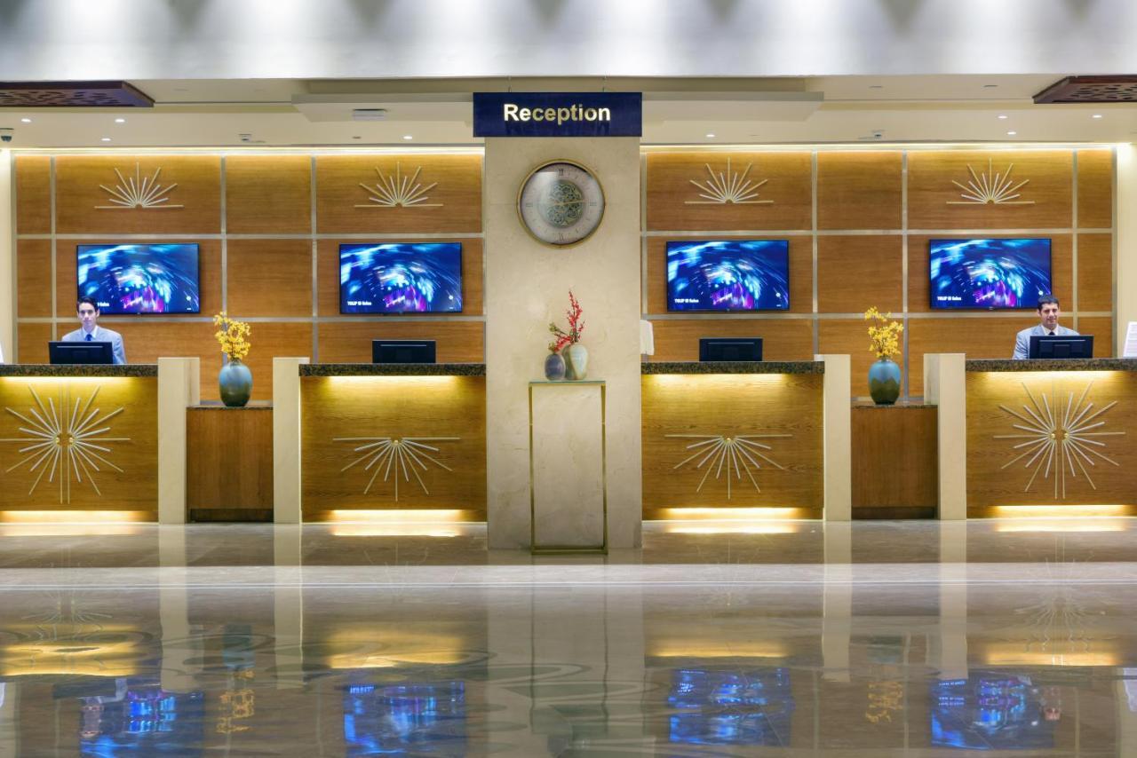 Tolip Resort El Galala Majestic Ajn Suchna Zewnętrze zdjęcie The image shows a hotel reception area. There are several reception desks lined up against a wall with decorative paneling. Each desk has a staff member, and above the desks, there are screens displaying information. A clock is visible on the wall, a