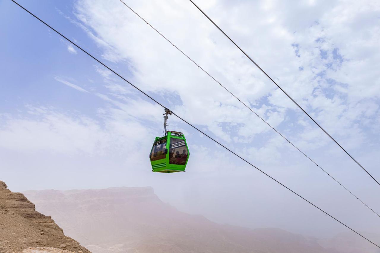 Tolip Resort El Galala Majestic Ajn Suchna Zewnętrze zdjęcie The photo shows a green cable car suspended in the air, traveling along overhead cables. The backdrop features a hazy landscape, likely mountainous or elevated terrain, with a cloudy sky. The scene suggests a scenic transportation method, possibly us