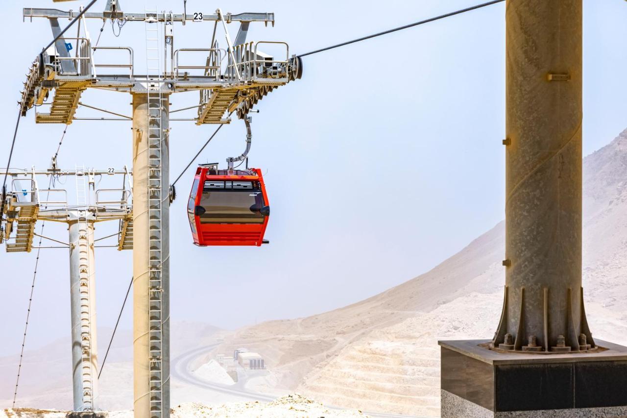 Tolip Resort El Galala Majestic Ajn Suchna Zewnętrze zdjęcie The photo shows a cable car system with a single red gondola suspended mid-air. It appears to be part of a larger cable car infrastructure, with metal pylons supporting the cables above. The background features a hazy landscape, likely characterized 