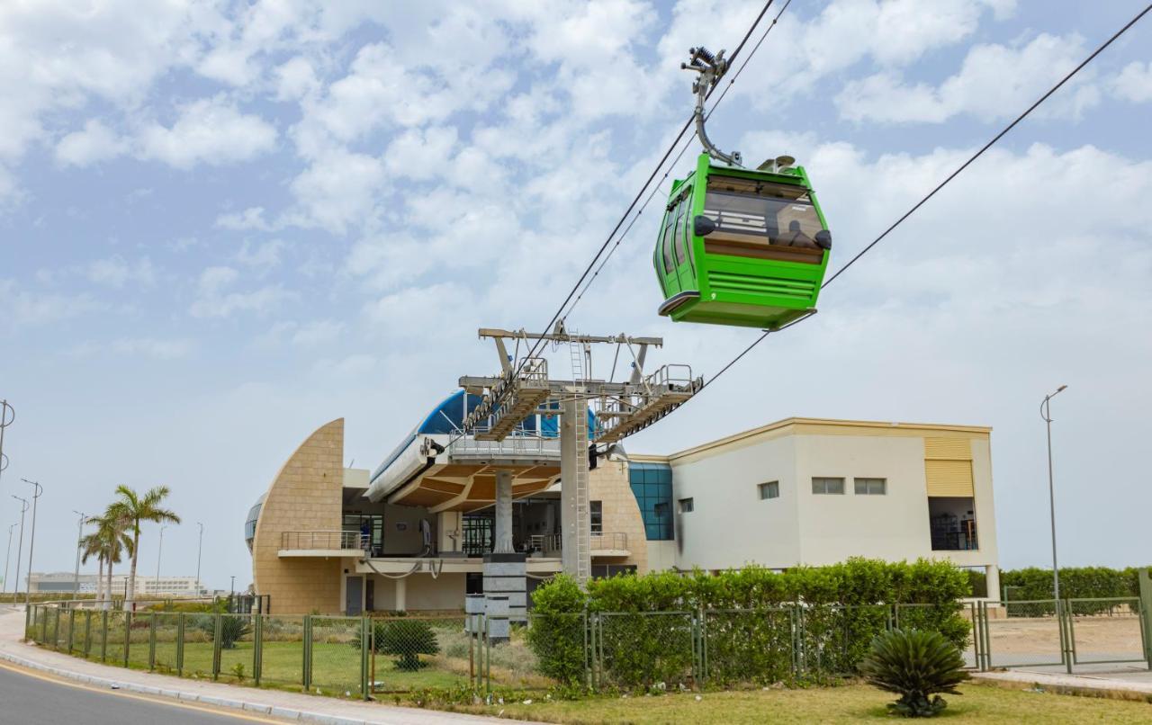 Tolip Resort El Galala Majestic Ajn Suchna Zewnętrze zdjęcie The photo shows a cable car or gondola system in operation, featuring a green-colored gondola. It is suspended from cables and moving towards a station, which appears to be a modern building with a unique architectural design. The station has large w