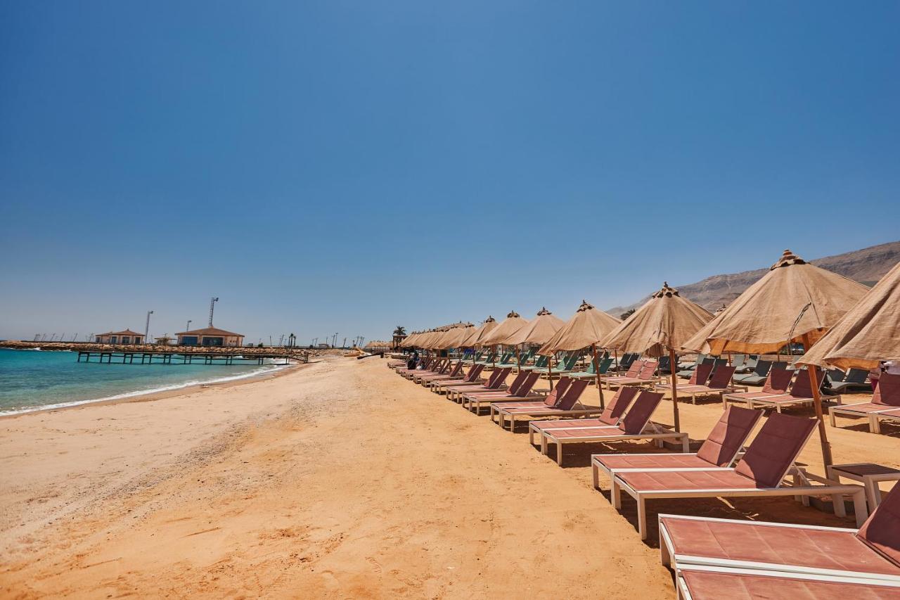 Tolip Resort El Galala Majestic Ajn Suchna Zewnętrze zdjęcie The photo shows a scenic beach with a stretch of golden sand under a clear blue sky. There are several beach loungers arranged in rows, each accompanied by thatched umbrellas providing shade. In the background, there is a wooden pier extending into t
