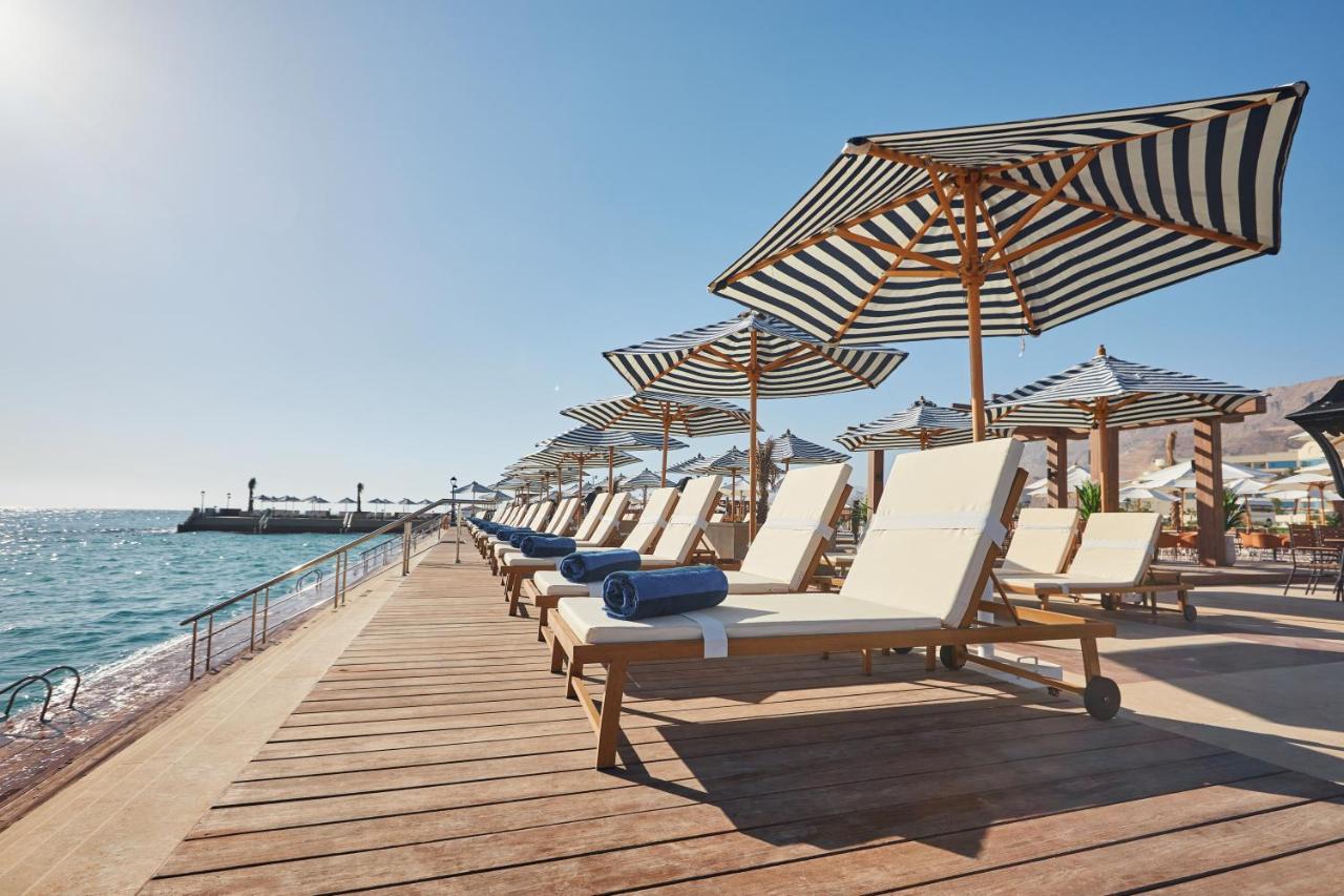 Tolip Resort El Galala Majestic Ajn Suchna Zewnętrze zdjęcie The photo depicts a serene beachside setting featuring a row of sun loungers arranged on a wooden deck. Each lounger is equipped with a folded towel and shaded by striped umbrellas. In the background, the water sparkles under bright sunlight, and the