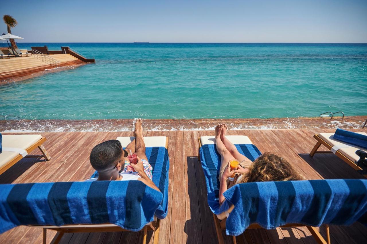 Tolip Resort El Galala Majestic Ajn Suchna Zewnętrze zdjęcie The photo depicts two people relaxing on lounge chairs by the waterfront. They are positioned on a wooden deck with a view of the clear, turquoise water. Both individuals are reclining, enjoying the sun, and each has a drink in hand. The scene convey