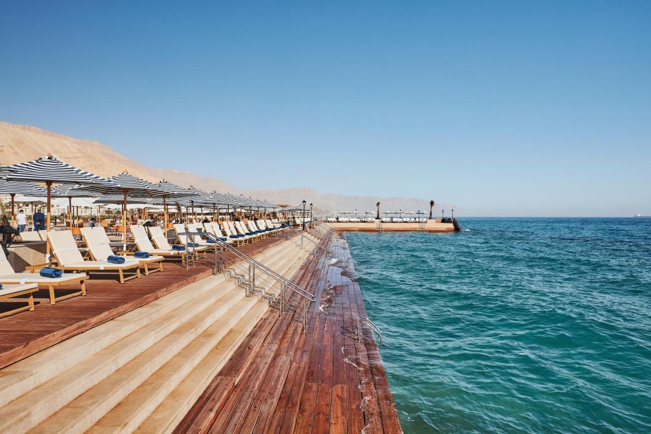 Tolip Resort El Galala Majestic Ajn Suchna Zewnętrze zdjęcie The photo shows a seaside area featuring a wooden deck that extends into the water. On the left side, there are several lounge chairs arranged neatly under shade structures, suggesting a relaxing atmosphere. The ocean is visible in the foreground, wi