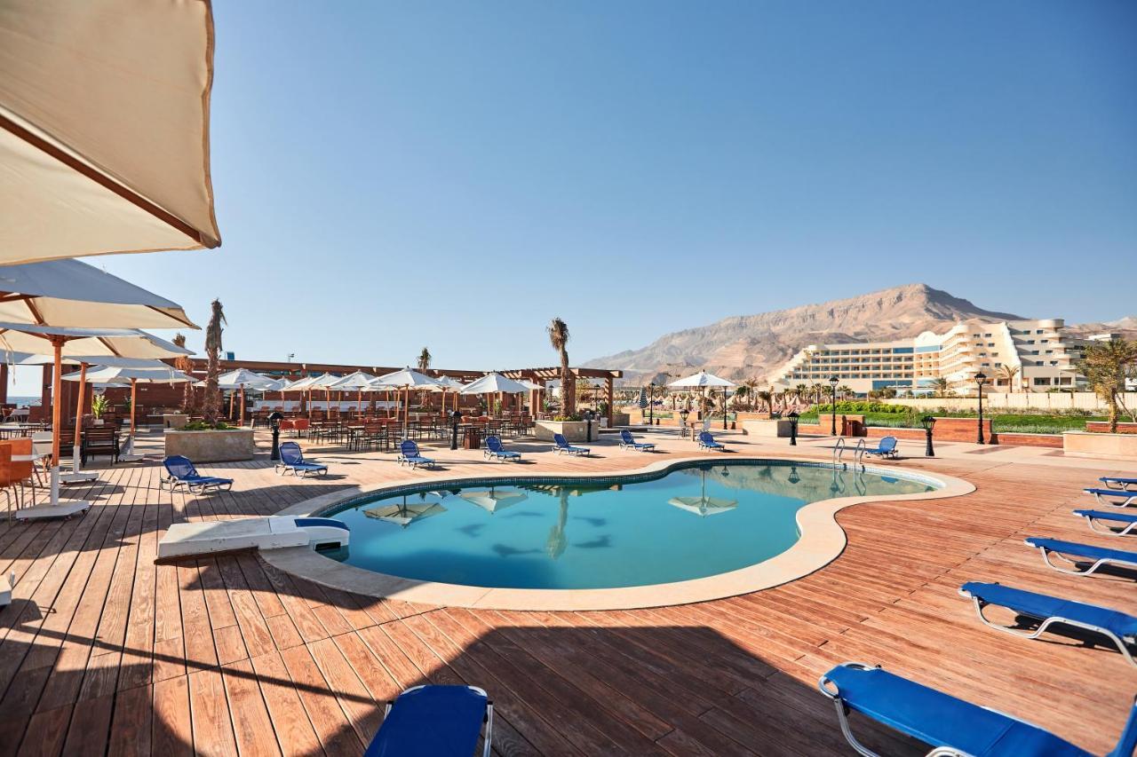 Tolip Resort El Galala Majestic Ajn Suchna Zewnętrze zdjęcie The photo shows a swimming pool area surrounded by sun loungers and umbrellas. The pool has a unique, curvy shape and is situated on a wooden deck. In the background, there are several buildings, likely part of a resort or hotel, and a mountainous la