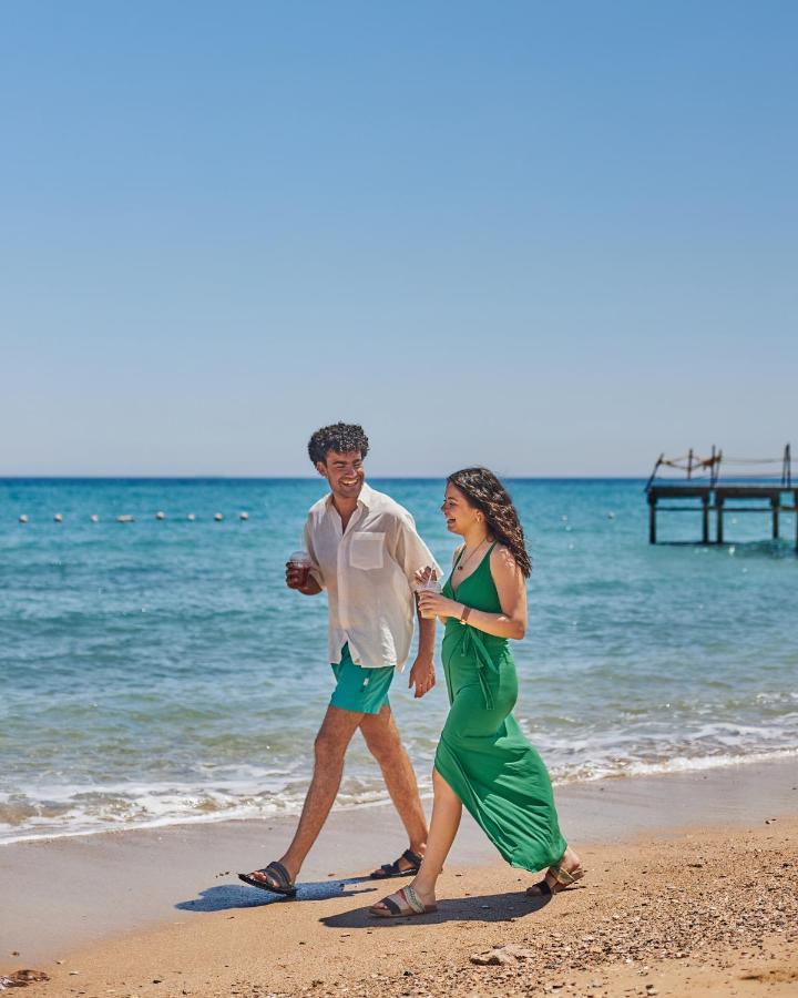 Tolip Resort El Galala Majestic Ajn Suchna Zewnętrze zdjęcie The photo shows a couple walking along a beach. The man is wearing a white short-sleeved shirt and green shorts, while the woman is dressed in a flowing green dress. They both appear to be enjoying the pleasant weather, with the ocean in the backgrou