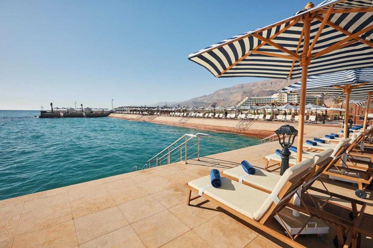 Tolip Resort El Galala Majestic Ajn Suchna Zewnętrze zdjęcie The photo shows a scenic beachside area with clear blue water. In the foreground, there are several sun loungers set up on a tiled surface, with blue towels neatly rolled on each one. The loungers are shaded by striped umbrellas. In the distance, a w