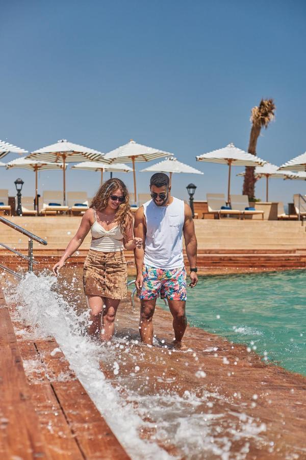 Tolip Resort El Galala Majestic Ajn Suchna Zewnętrze zdjęcie The photo depicts a couple walking along a poolside area. They are enjoying a sunny day near a swimming pool, with splashes of water around them. The woman is wearing a light-colored top and a patterned skirt, while the man is dressed in a sleeveless