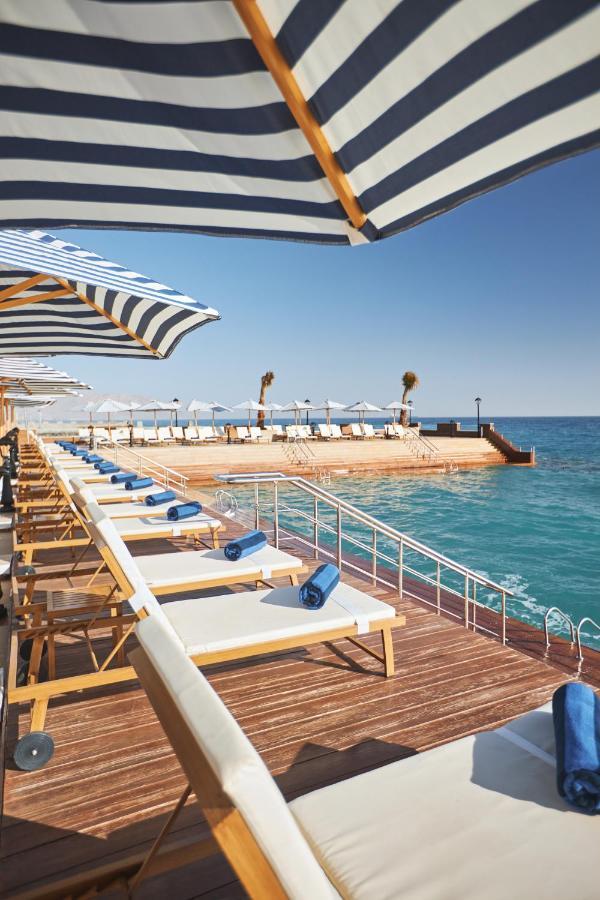 Tolip Resort El Galala Majestic Ajn Suchna Zewnętrze zdjęcie The photo depicts a serene outdoor area by the waterfront. There are several lounge chairs arranged in a row, each equipped with blue towels. Large, striped umbrellas provide shade over some of the chairs. In the background, there is a wooden deck th