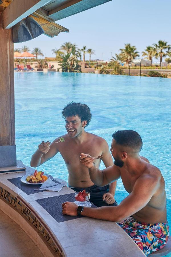 Tolip Resort El Galala Majestic Ajn Suchna Zewnętrze zdjęcie The photo features two young men enjoying a meal together. They are sitting at a poolside table with a clear blue swimming pool in the background. The men are shirtless, suggesting a casual, relaxed atmosphere, likely in a warm environment. One man i