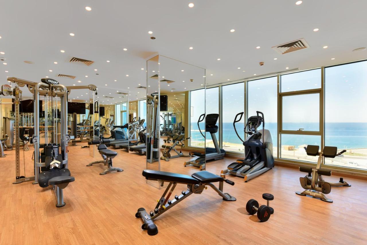 Tolip Resort El Galala Majestic Ajn Suchna Zewnętrze zdjęcie The photo shows a modern gym interior with a variety of fitness equipment. There are cardio machines like treadmills and ellipticals, along with strength training equipment such as free weights and benches. The gym features large windows that offer a