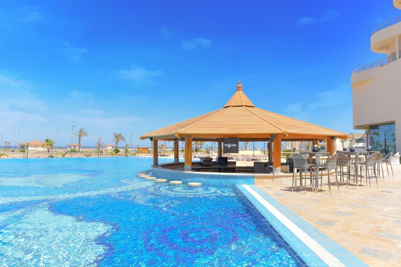 Tolip Resort El Galala Majestic Ajn Suchna Zewnętrze zdjęcie The photo shows a beautiful outdoor setting featuring a large swimming pool with clear blue water. In the center, there is a thatched-roof gazebo, which serves as a shaded seating area. Surrounding the gazebo are lounge chairs and a bar area with hig