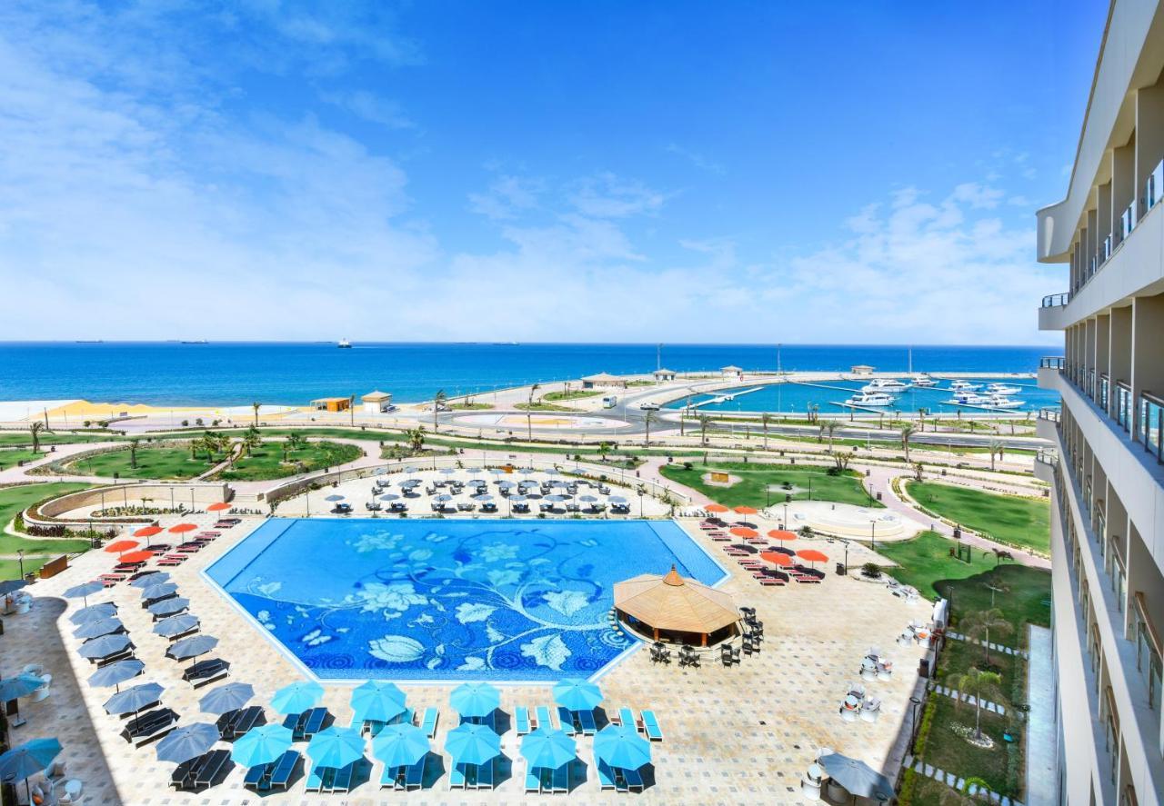 Tolip Resort El Galala Majestic Ajn Suchna Zewnętrze zdjęcie The photo shows a beautiful outdoor view of a resort area featuring a large swimming pool with a decorative underwater design. Surrounding the pool are numerous sun loungers and umbrellas in a variety of colors. In the background, there is a scenic c