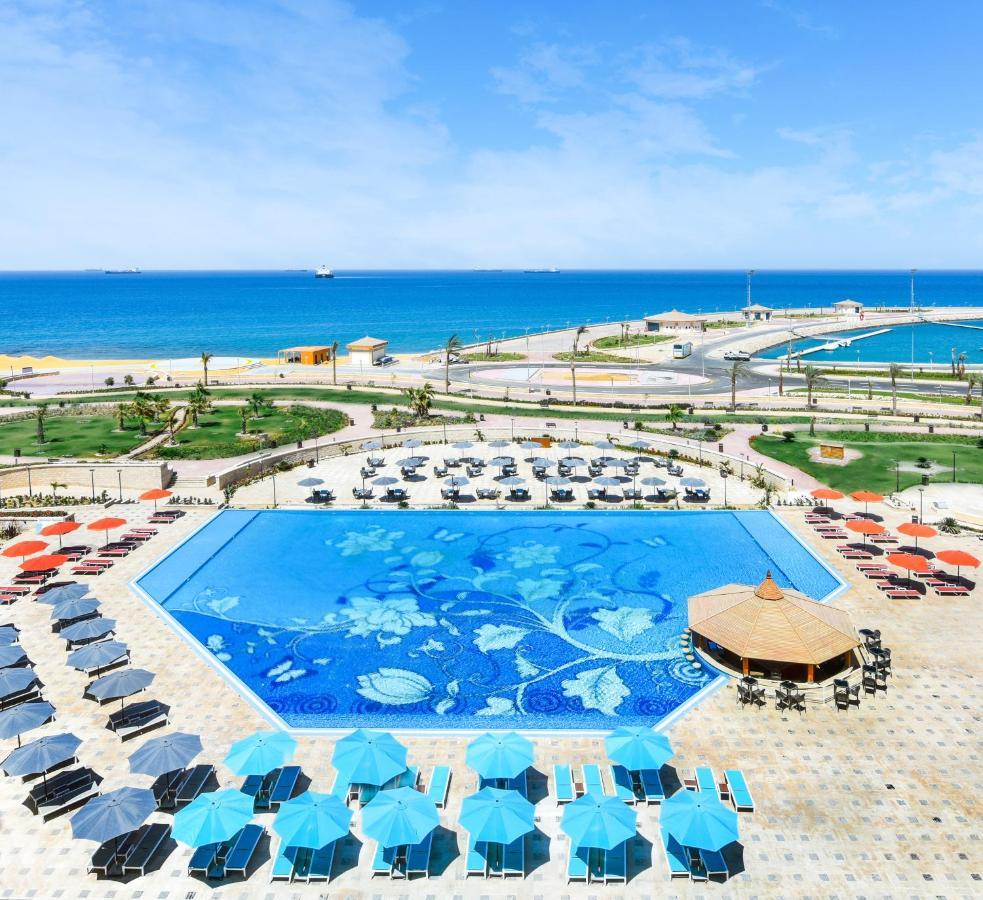 Tolip Resort El Galala Majestic Ajn Suchna Zewnętrze zdjęcie The photo features a large swimming pool with a decorative design on its surface, surrounded by sun loungers and umbrellas. In the background, there is a view of the sea, with a clear blue sky and a few scattered clouds. The area is well-maintained, 