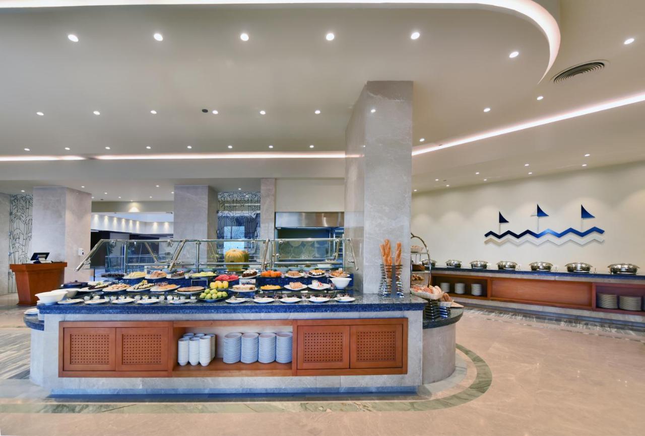 Tolip Resort El Galala Majestic Ajn Suchna Zewnętrze zdjęcie The image shows a buffet-style dining area in a restaurant or hotel. There are several food stations with a variety of dishes displayed, including fruits, salads, and other food items. The layout features a large, central island with a wooden base an