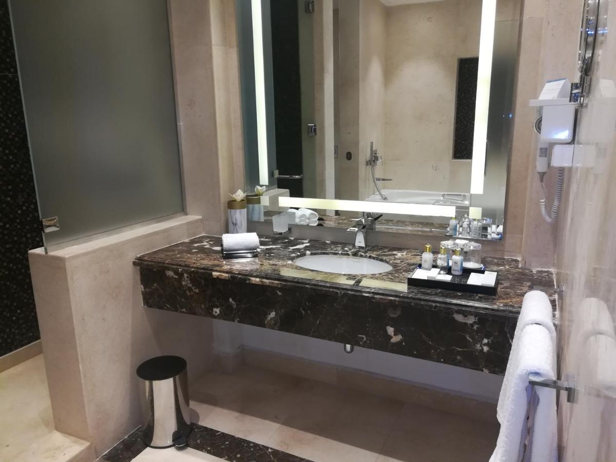 Tolip Resort El Galala Majestic Ajn Suchna Zewnętrze zdjęcie The photo shows a modern bathroom with elegant design features. There is a large vanity made of dark marble with a sink and a set of toiletries neatly arranged on top. A mirror with built-in lighting is mounted above the sink. To the side, there is a