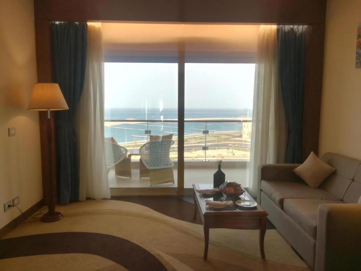 Tolip Resort El Galala Majestic Ajn Suchna Zewnętrze zdjęcie The photo shows a stylish interior of a room with a large window overlooking the sea. The window features sheer and thick curtains and is framed by a balcony with two chairs visible outside. Inside, there is a comfortable-looking sofa, a coffee table