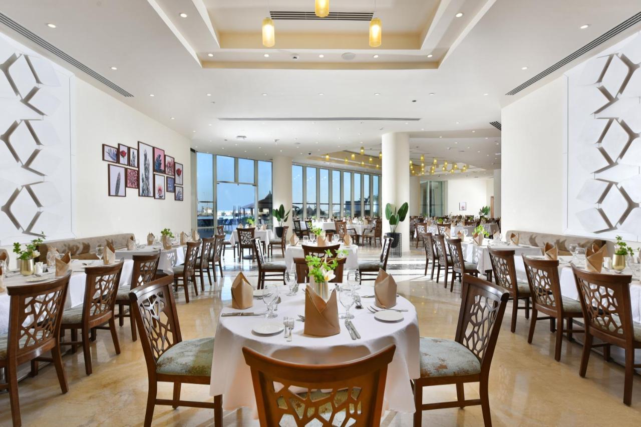 Tolip Resort El Galala Majestic Ajn Suchna Zewnętrze zdjęcie The photo displays a spacious and elegantly designed restaurant interior. It features numerous tables set for dining, with white tablecloths, neatly folded napkins, and decorative centerpieces of flowers. The seating consists of wooden chairs that co